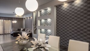 A modern living room with a glass coffee table, white chairs, and a sofa. There are pendant lights and built-in shelves with decorative items. - Callista Luxury Residences (Ierapetra)