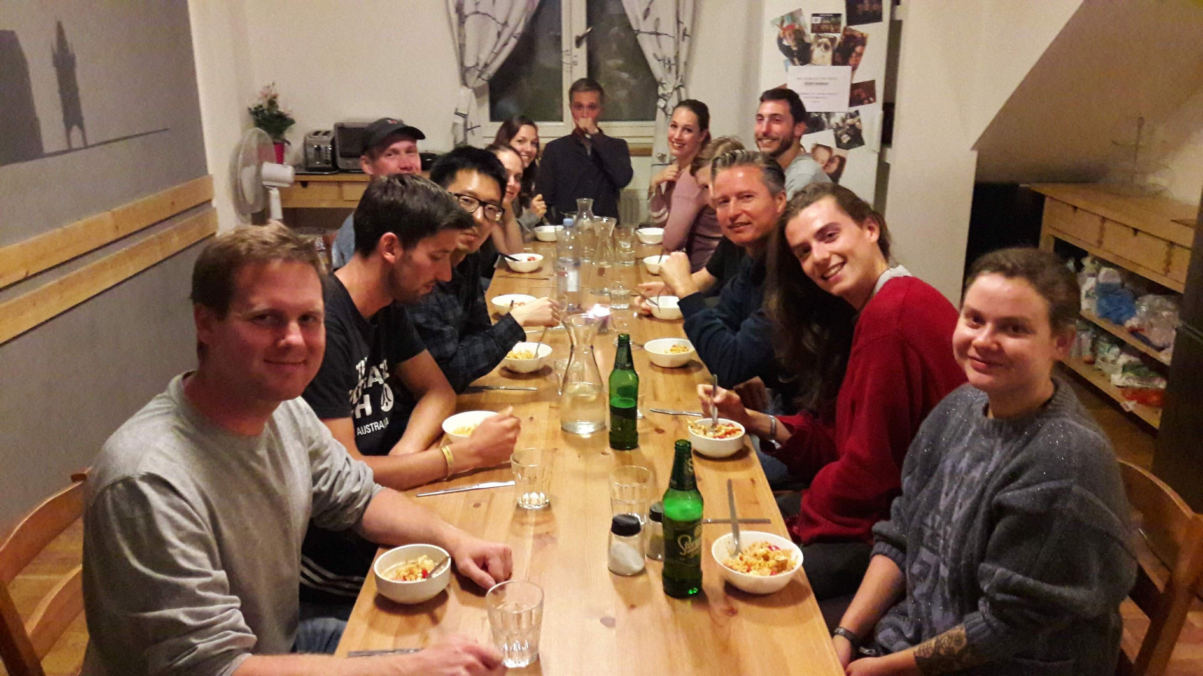 Un groupe de personnes est assis autour d’une longue table en bois, devant des bols remplis de nourriture, des verres et des bouteilles, dans une pièce dotée d’une fenêtre et d’un mur orné de tableaux.