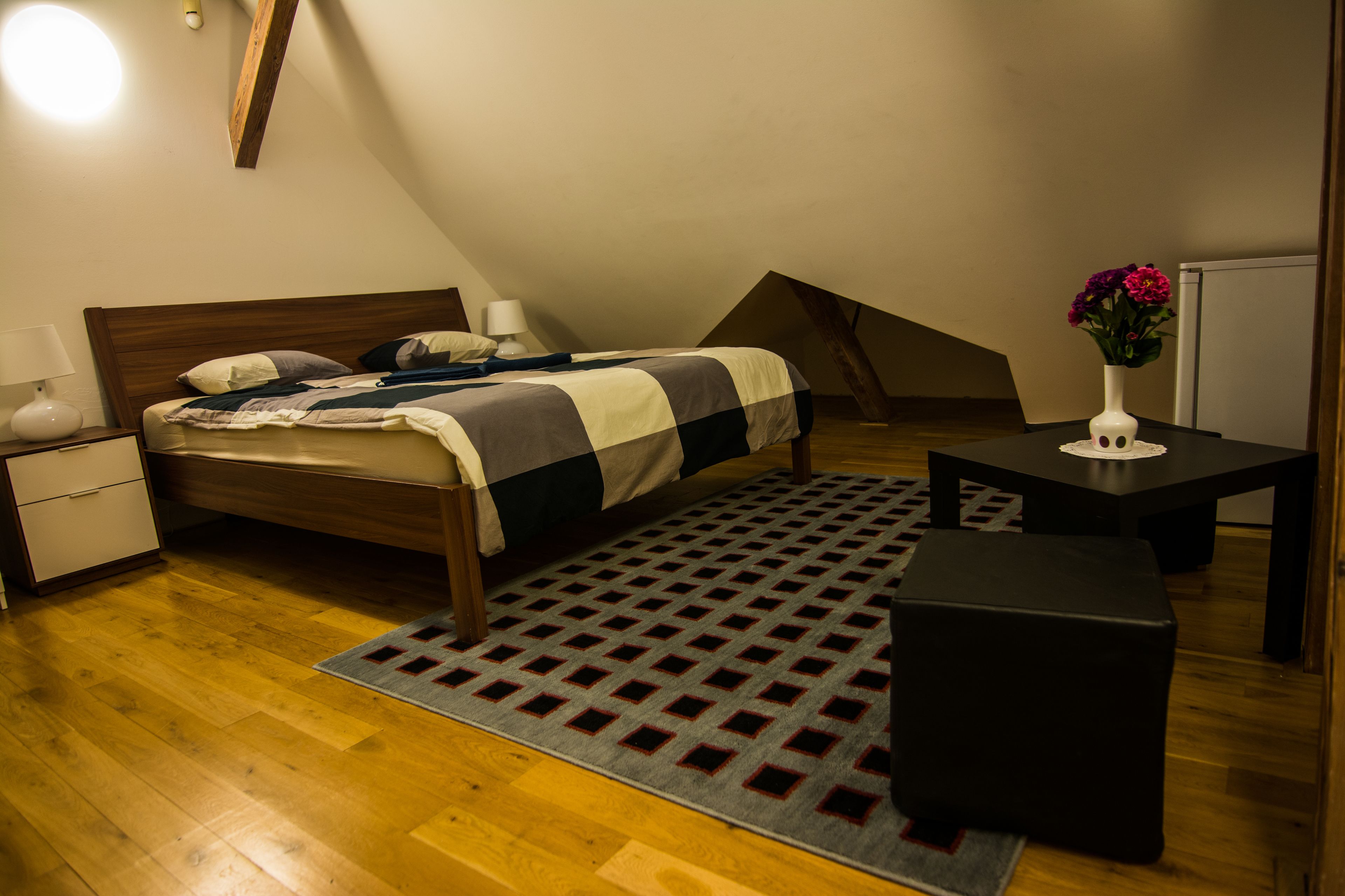 A bedroom with a wooden bed, a nightstand, a table with a vase of flowers, and a patterned rug.