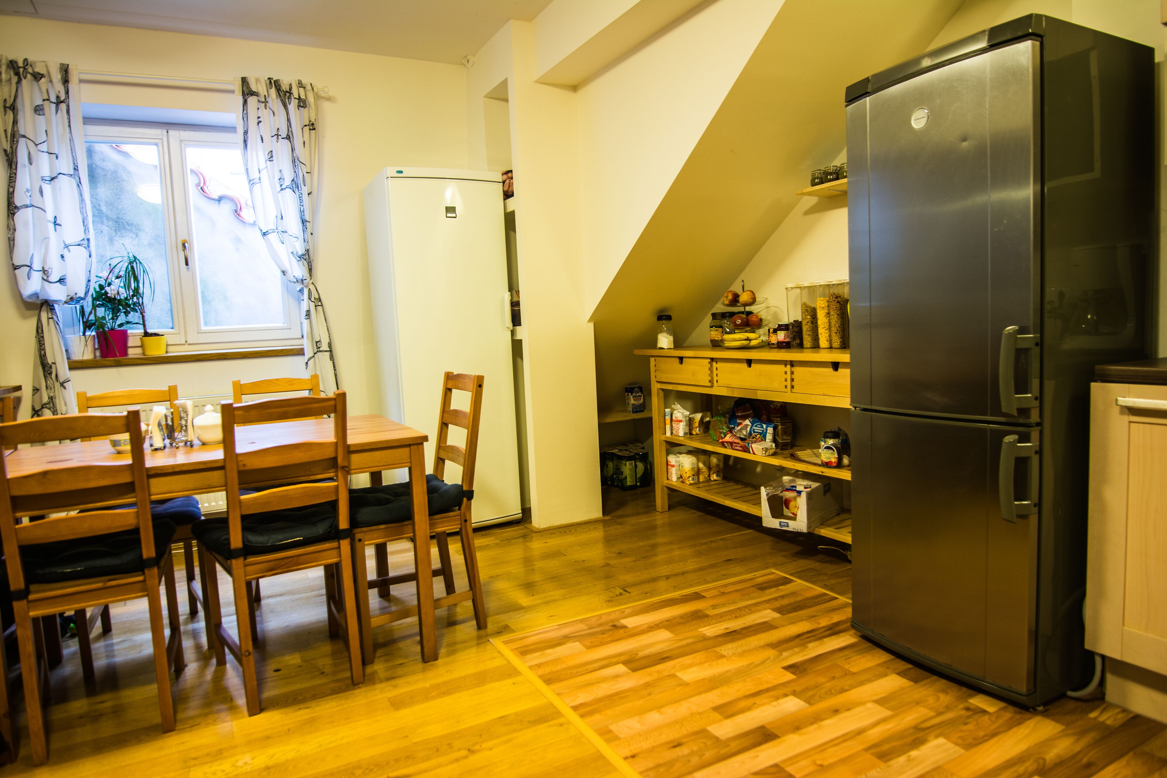 Une cuisine équipée d’une table à manger, de chaises, d’un réfrigérateur, d’étagères en bois et d’une fenêtre avec des rideaux.