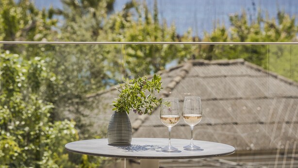 A table with a vase and two glasses of white wine, set against a backdrop of greenery and a view of the sea. - Notos Luxury Studios (Kefalonia)