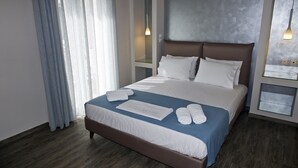 A neatly made bed with white linens, a blue accent on the bedspread, and a white towel with "NOLOS" on it. - Notos Luxury Studios (Kefalonia)