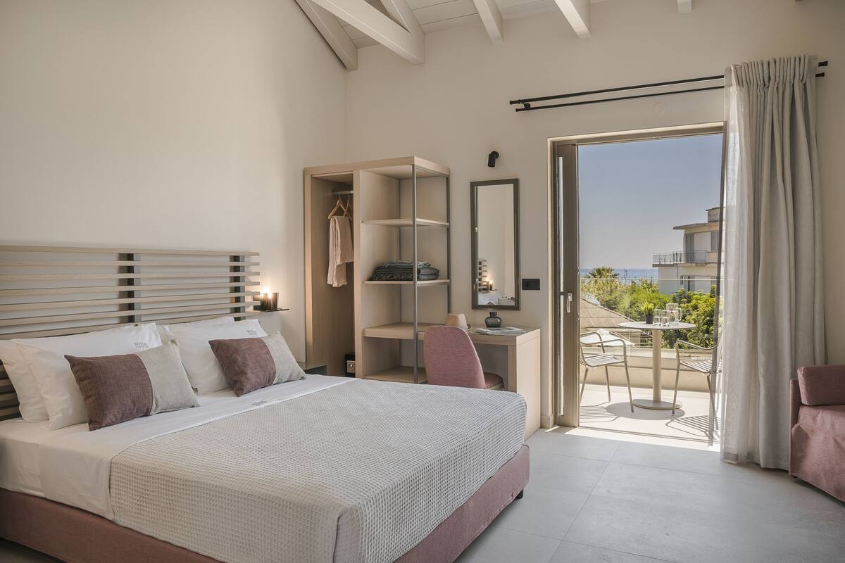 A bedroom with a bed, a desk, a chair, a wardrobe, and a balcony with a table and chairs.