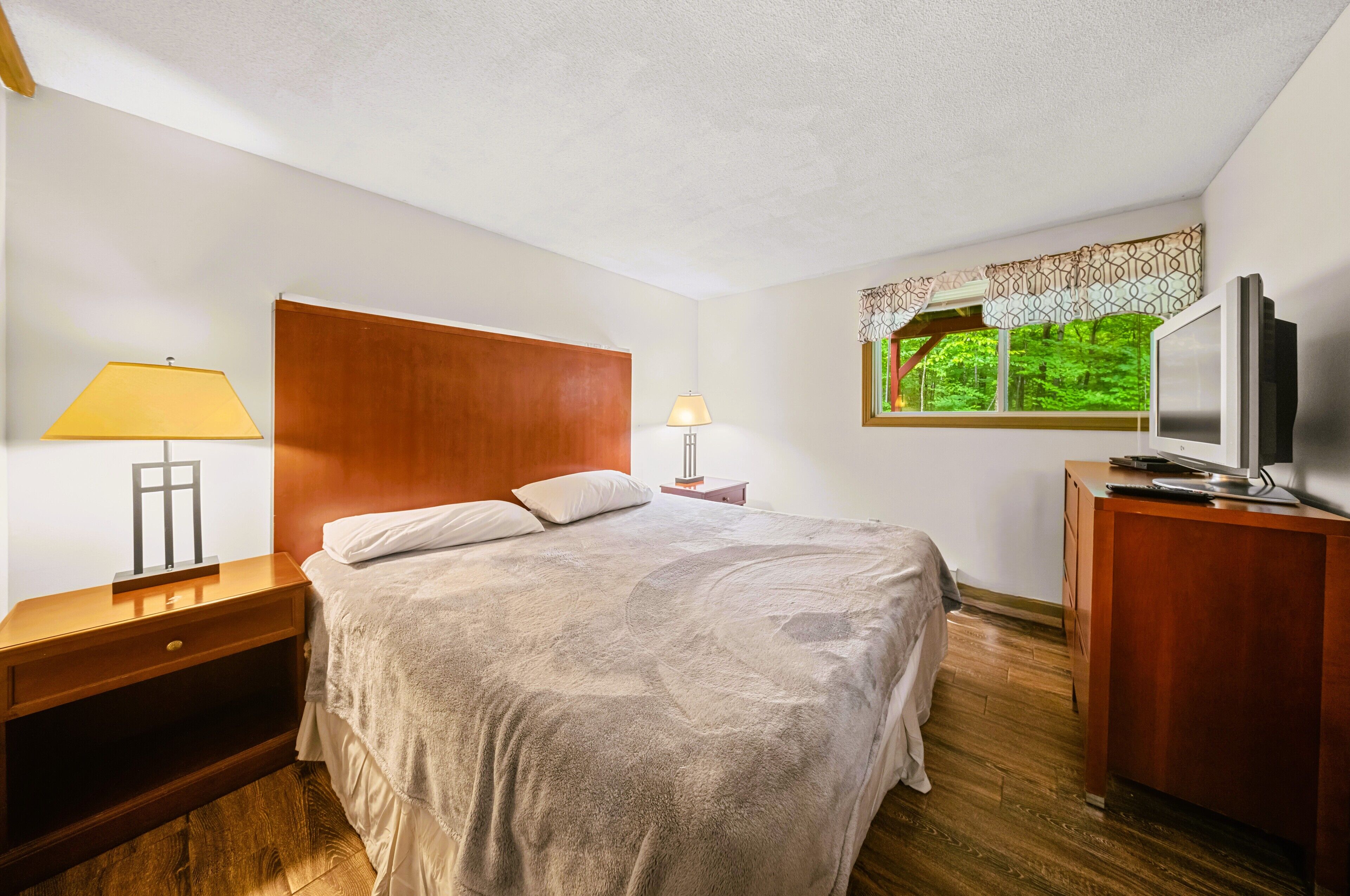 A bedroom with a large bed, wooden headboard, bedside lamps, a TV, and a window with a view of greenery.