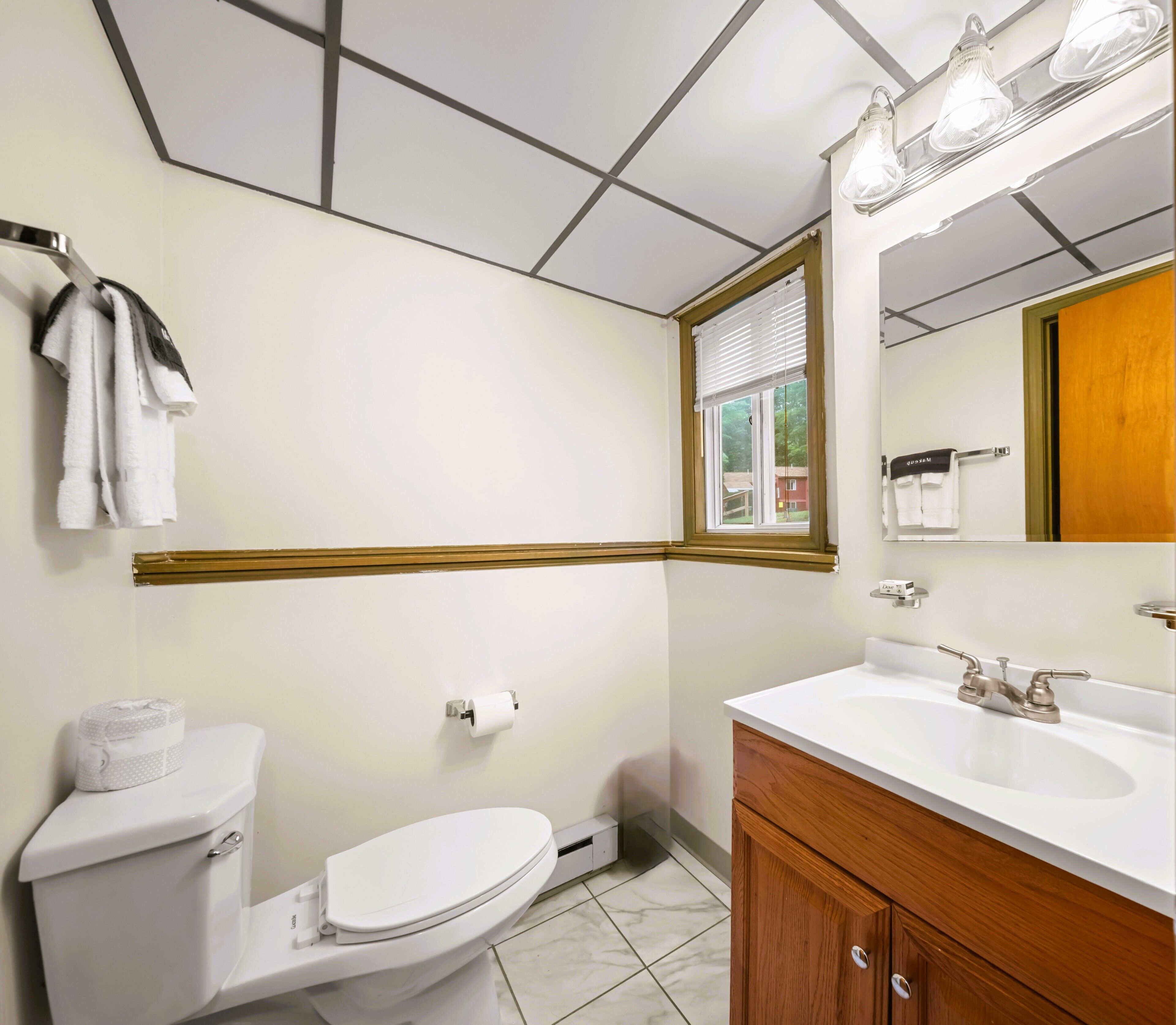 A bathroom with a toilet, sink, mirror, and window.
