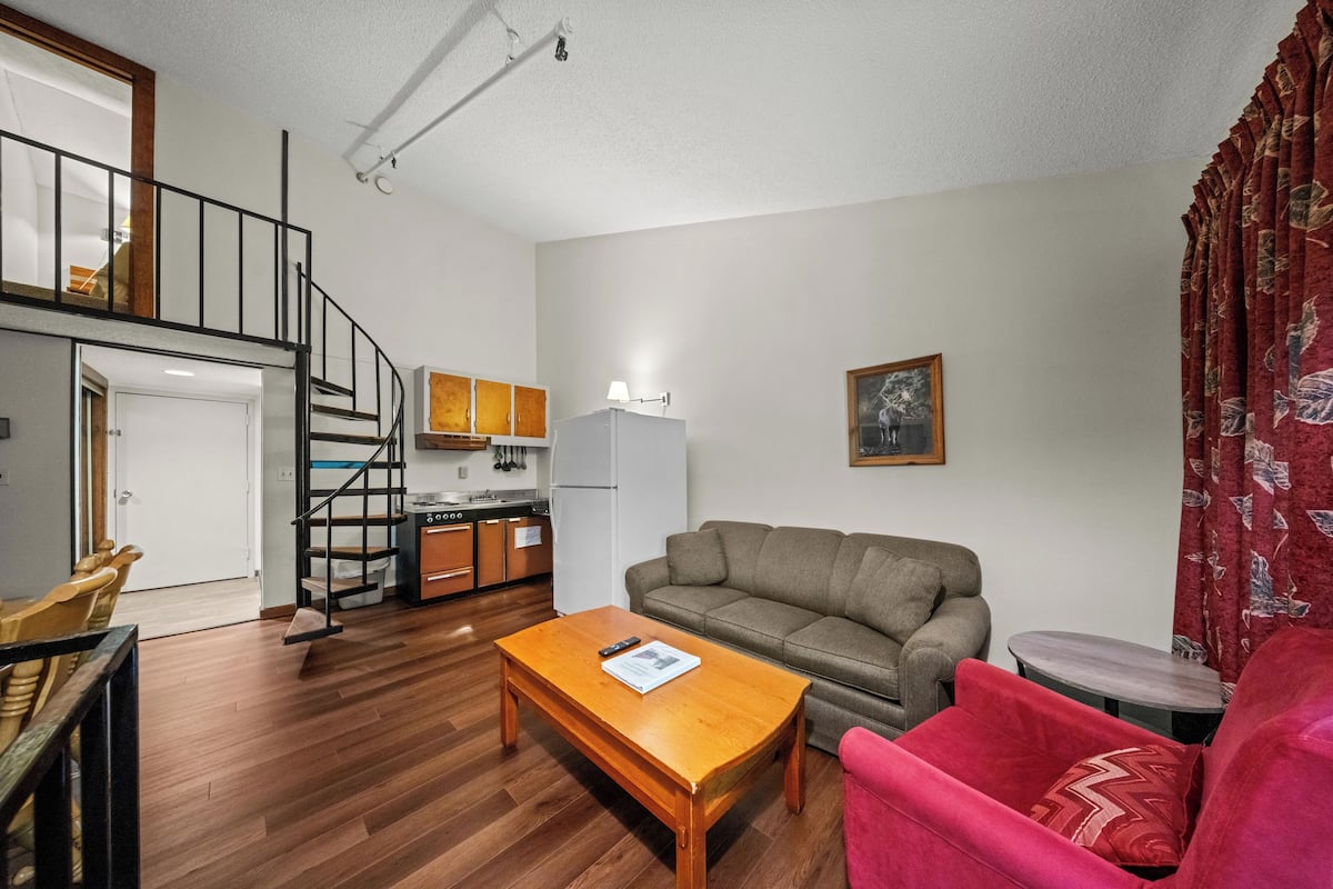 Um espaço compacto com uma escada que leva a um mezanino, uma cozinha compacta, um sofá, uma mesa de centro e uma poltrona vermelha.