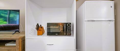A compact living space with a flat-screen TV, a microwave, a refrigerator, and a wooden desk.