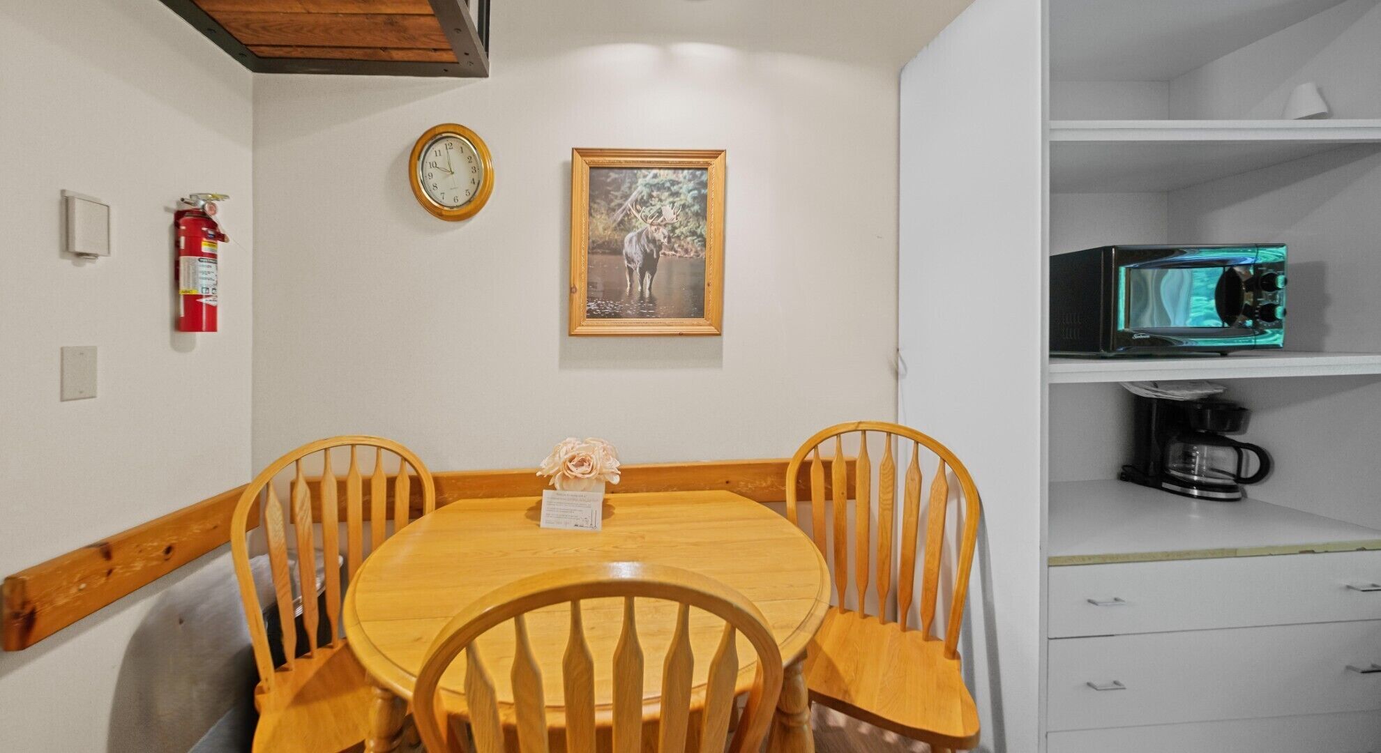 A dining area with a round wooden table and four chairs, a clock, a framed wildlife photograph, and a coffee machine on a white cabinet.