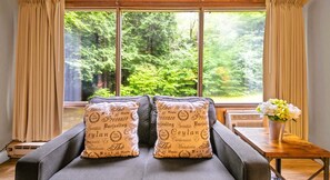 A room with a sofa facing a large window with a forest view, two decorative pillows with text, a small table with a vase of flowers, and a radiator.