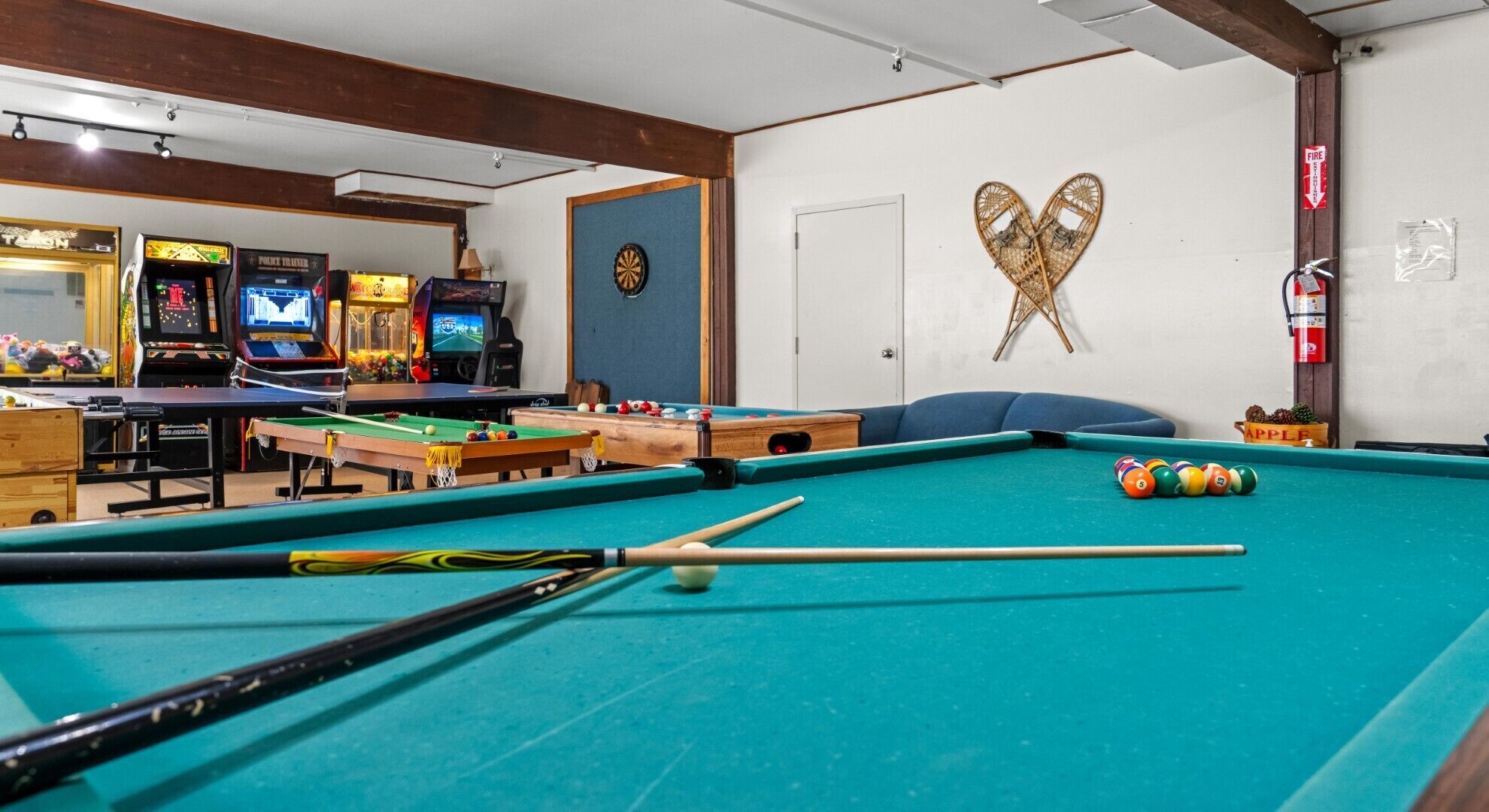 A pool table with billiard balls and cues, a dartboard, and arcade games in the background.