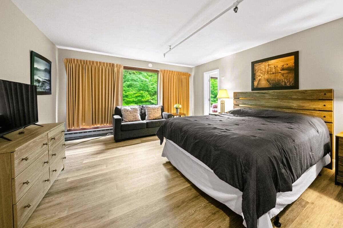 Quarto moderno com uma cama grande, uma televisão, uma cômoda de madeira e vista para o verde pela janela.