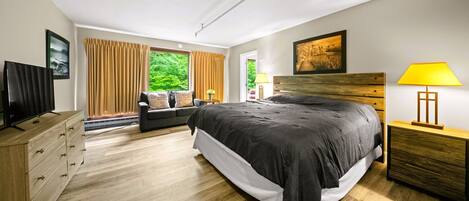 A modern bedroom with a large bed, a TV, a wooden dresser, and a view of greenery through the window.