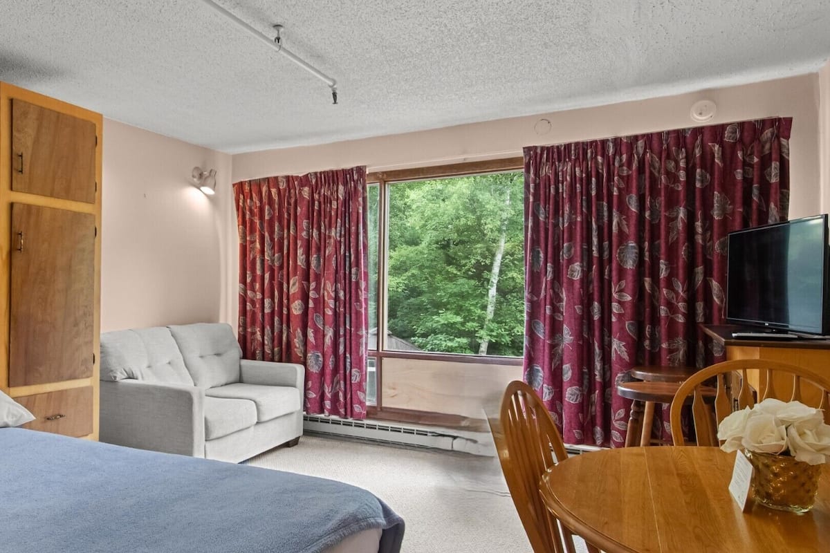 Quarto com cama, uma cadeira, escrivaninha com televisão e janela com cortinas.