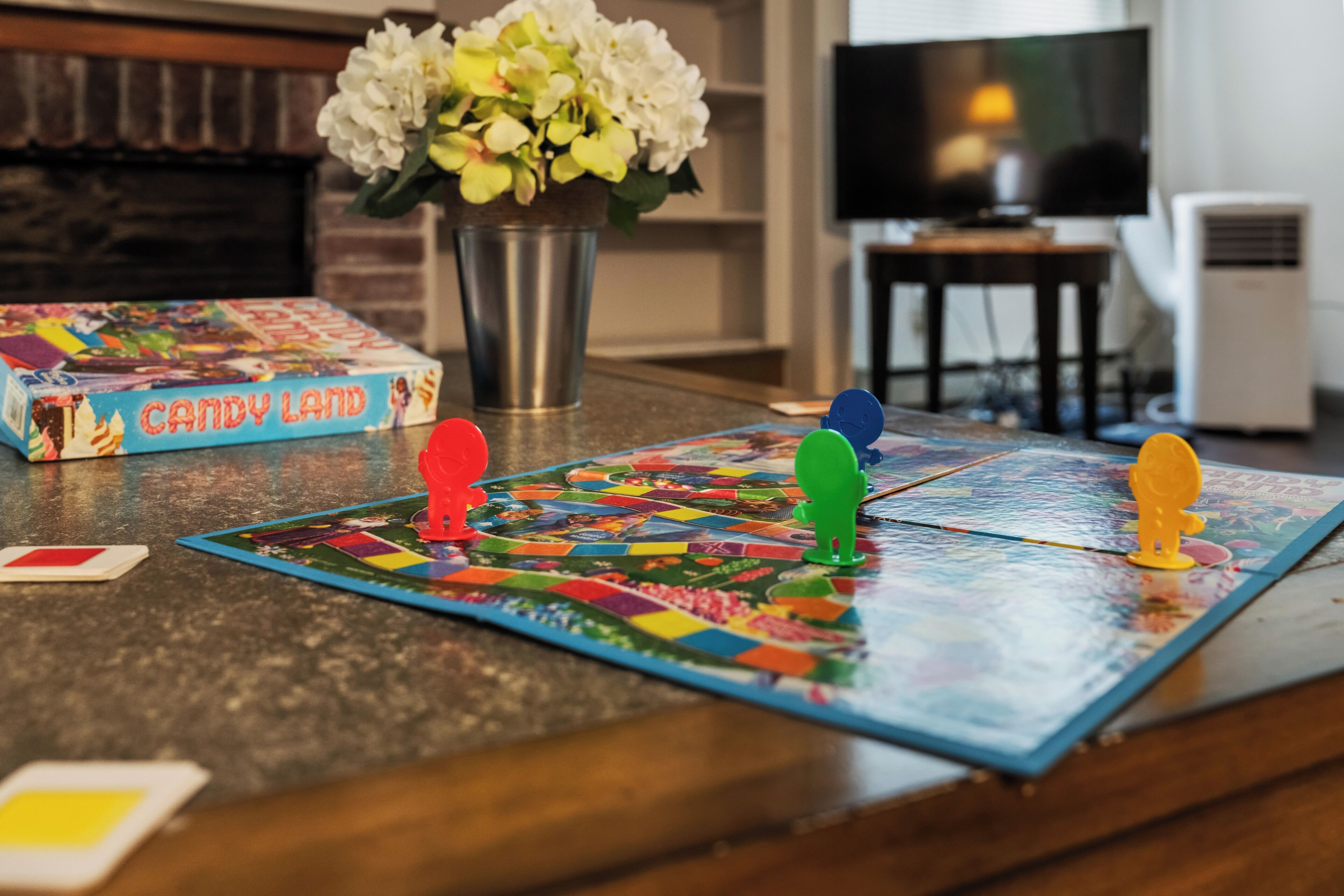 Un jeu de société est installé sur un comptoir de cuisine, avec un vase de fleurs et un téléviseur en arrière-plan.