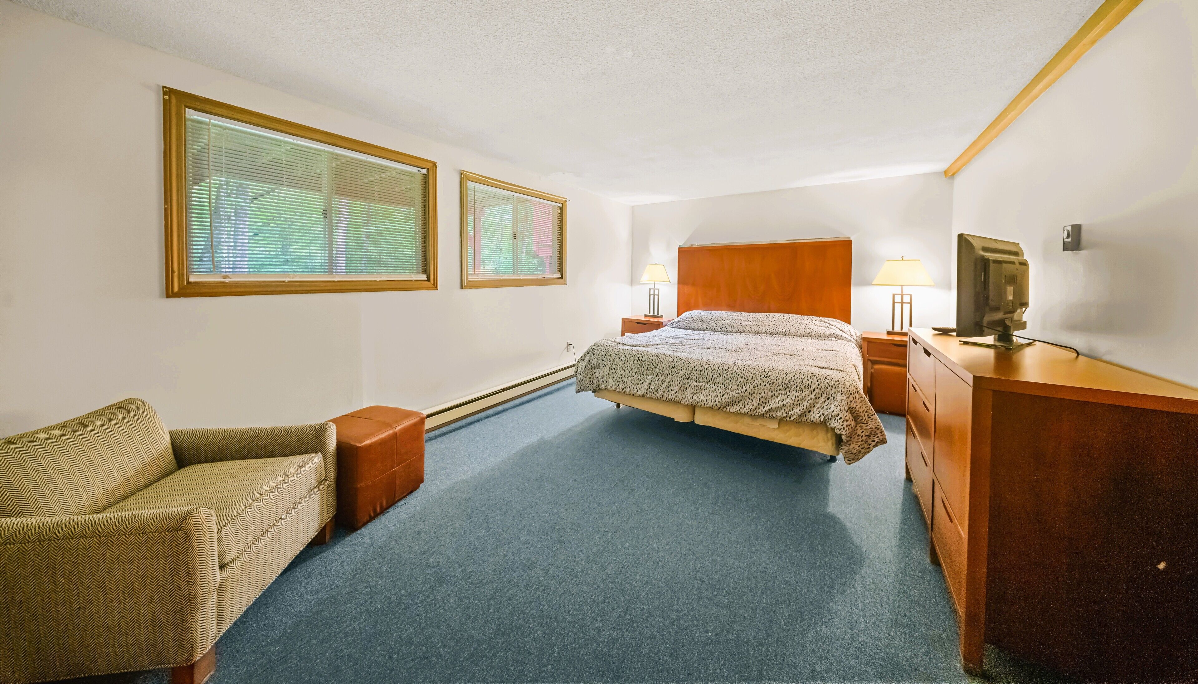 A hotel room with a bed, two bedside tables with lamps, a chair, a desk with a computer, and two windows with blinds.