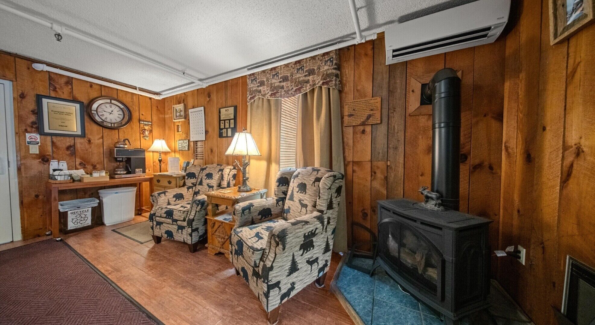 Una habitación acogedora con paredes de madera, una chimenea, dos sillones estampados, una mesita con una lámpara y un reloj en la pared.
