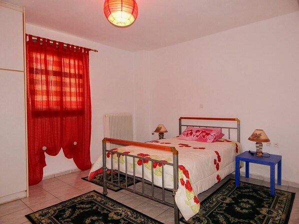 A bedroom with a metal bed frame, two bedside tables, a red curtain, a radiator, and a patterned rug. - Tripoli Apartments & Rooms (Tripoli)