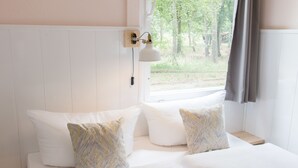 Ein Schlafzimmer mit einem Bett, zwei Kissen, einem Nachttisch, einer Lampe und einem Fenster mit Blick auf Bäume.