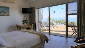 Ein Hotelzimmer mit Bett, Fernseher, Schreibtisch mit Pflanze und Balkon mit Stühlen und Meerblick.