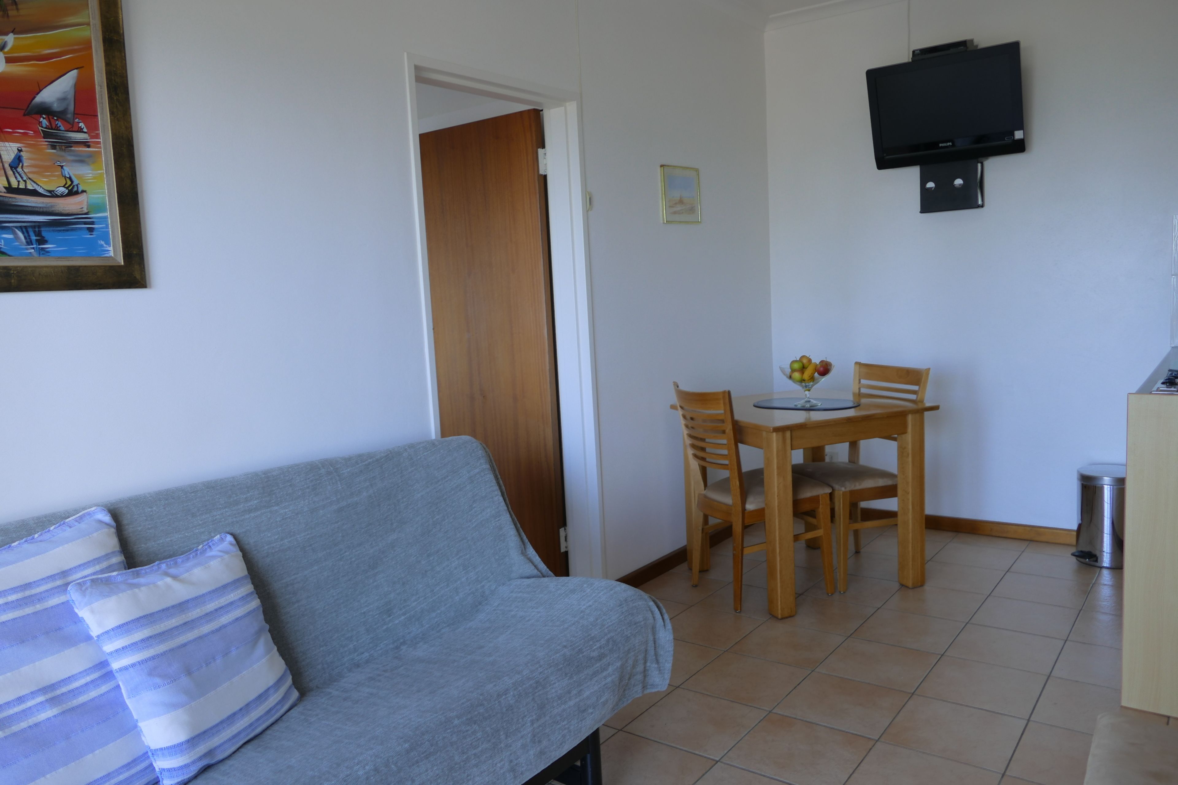 A room with a grey sofa, a wooden dining table with chairs, a TV mounted on the wall, and a door leading to another room.