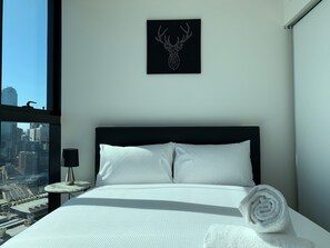 A modern hotel room with a large bed, white bedding, a city view, a framed deer head artwork, and a neatly made towel. - MYROOM at ShadowPlay (Southbank)