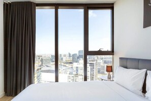 A hotel room with a large bed, a nightstand with a lamp, and a view of a cityscape through floor-to-ceiling windows. - MYROOM at ShadowPlay (Southbank)