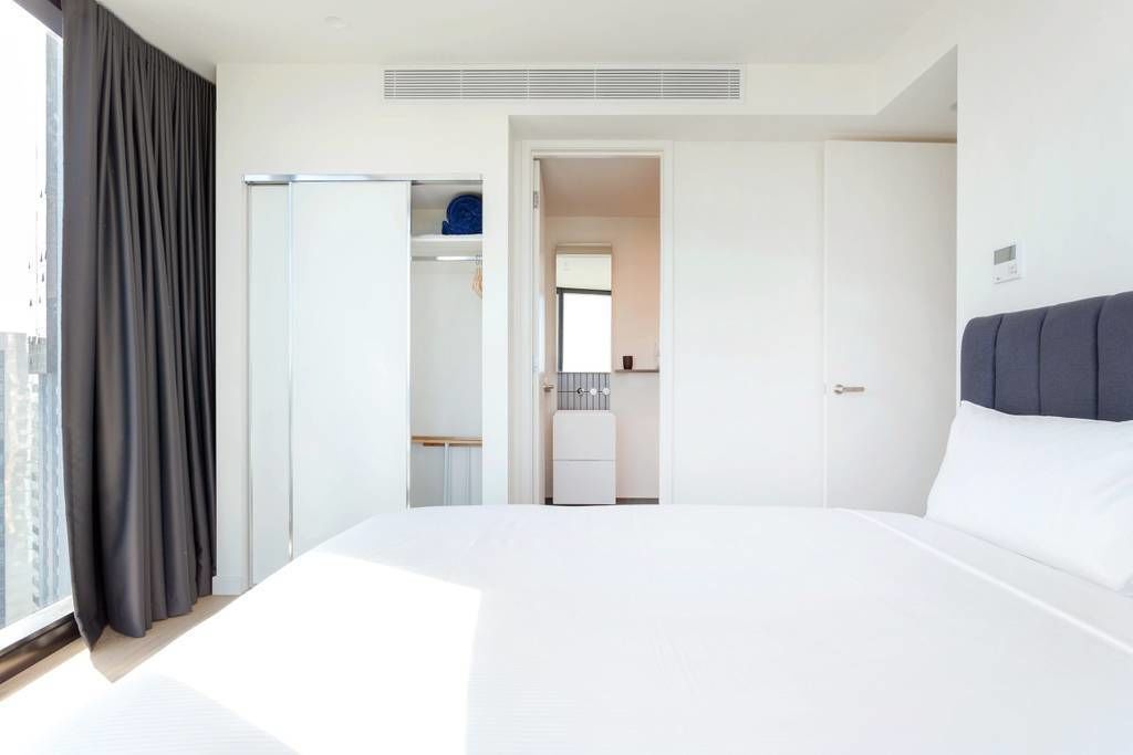 A hotel room with a large bed, a wardrobe, a kitchenette, and a view of the city.