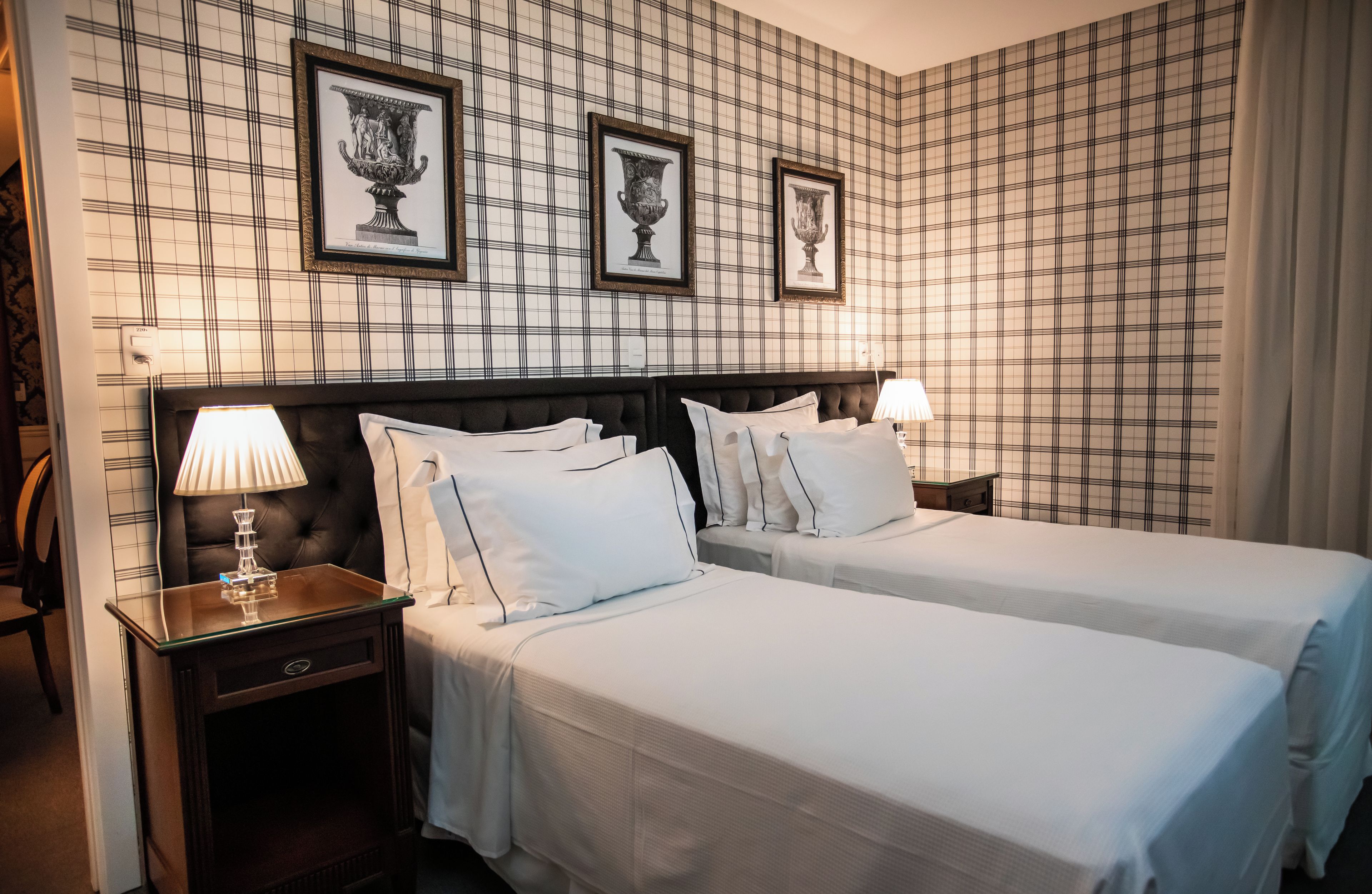 Habitación de hotel con un estampado de cuadros en la pared, dos camas con sábanas blancas, un cabecero oscuro y una mesita de noche con una lámpara.