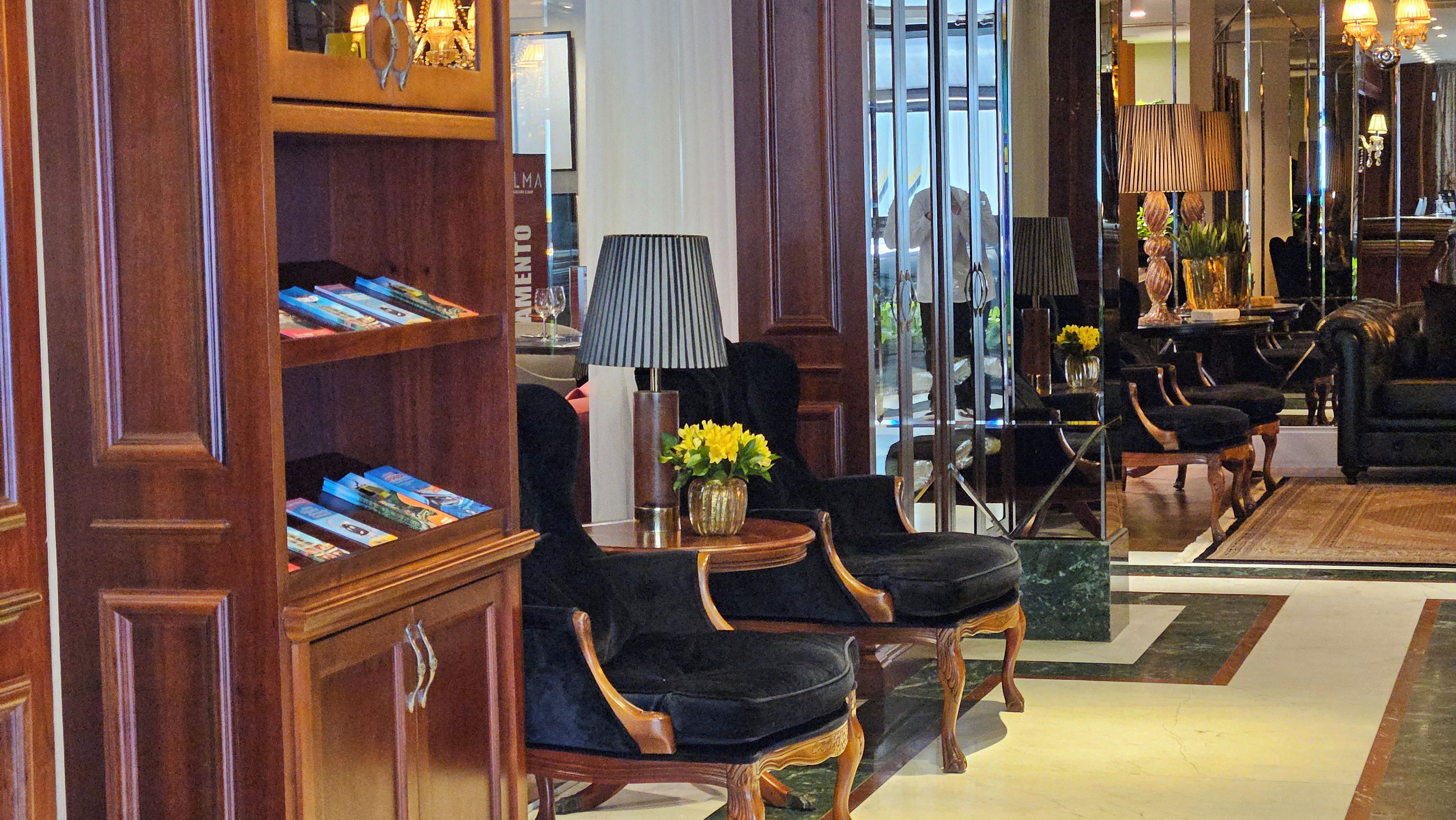 Een hal met een houten boekenkast, een zwarte beklede stoel, een lamp en een vaas met gele bloemen.