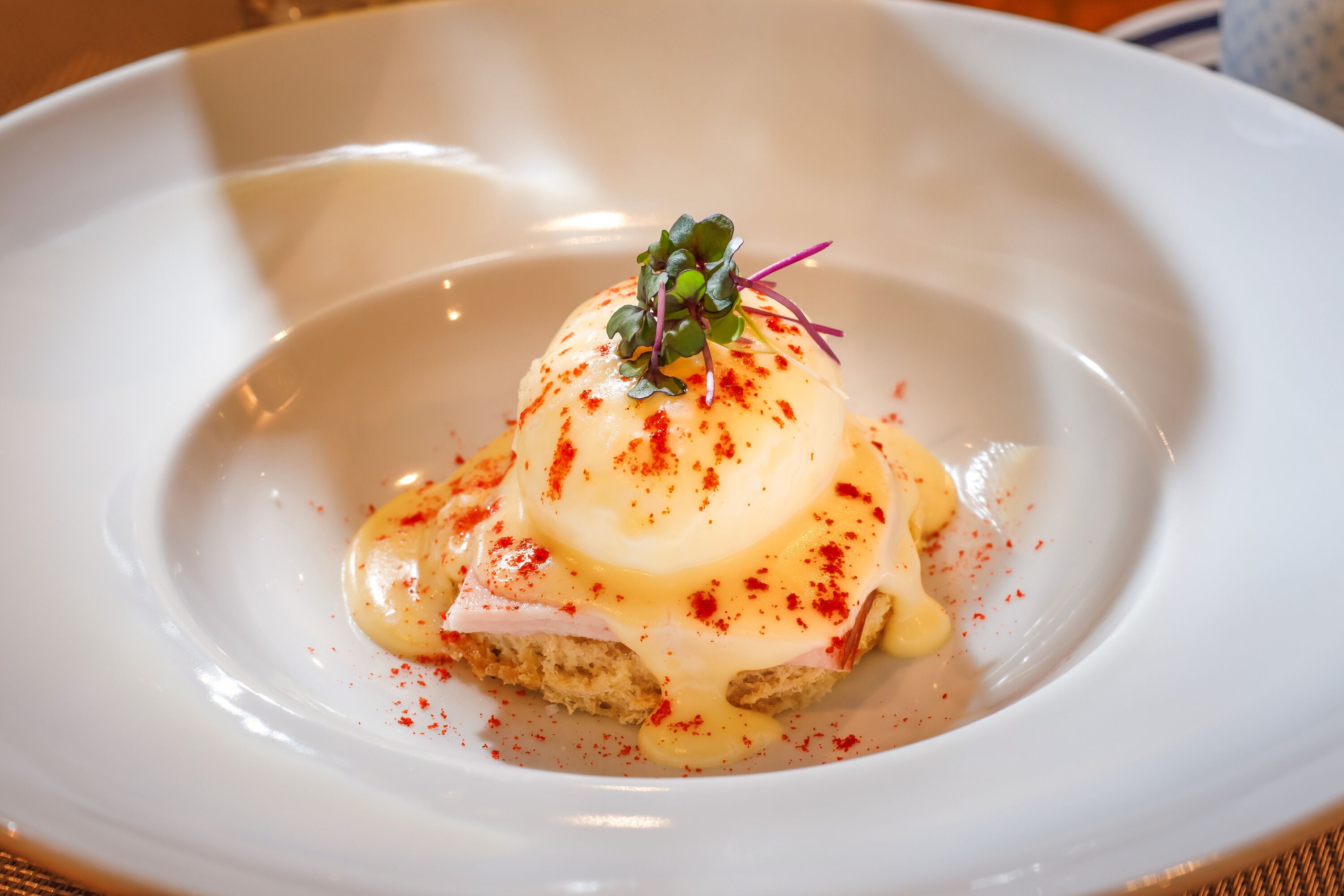 A white plate with a dish consisting of a poached egg, a slice of ham, and a garnish of microgreens.