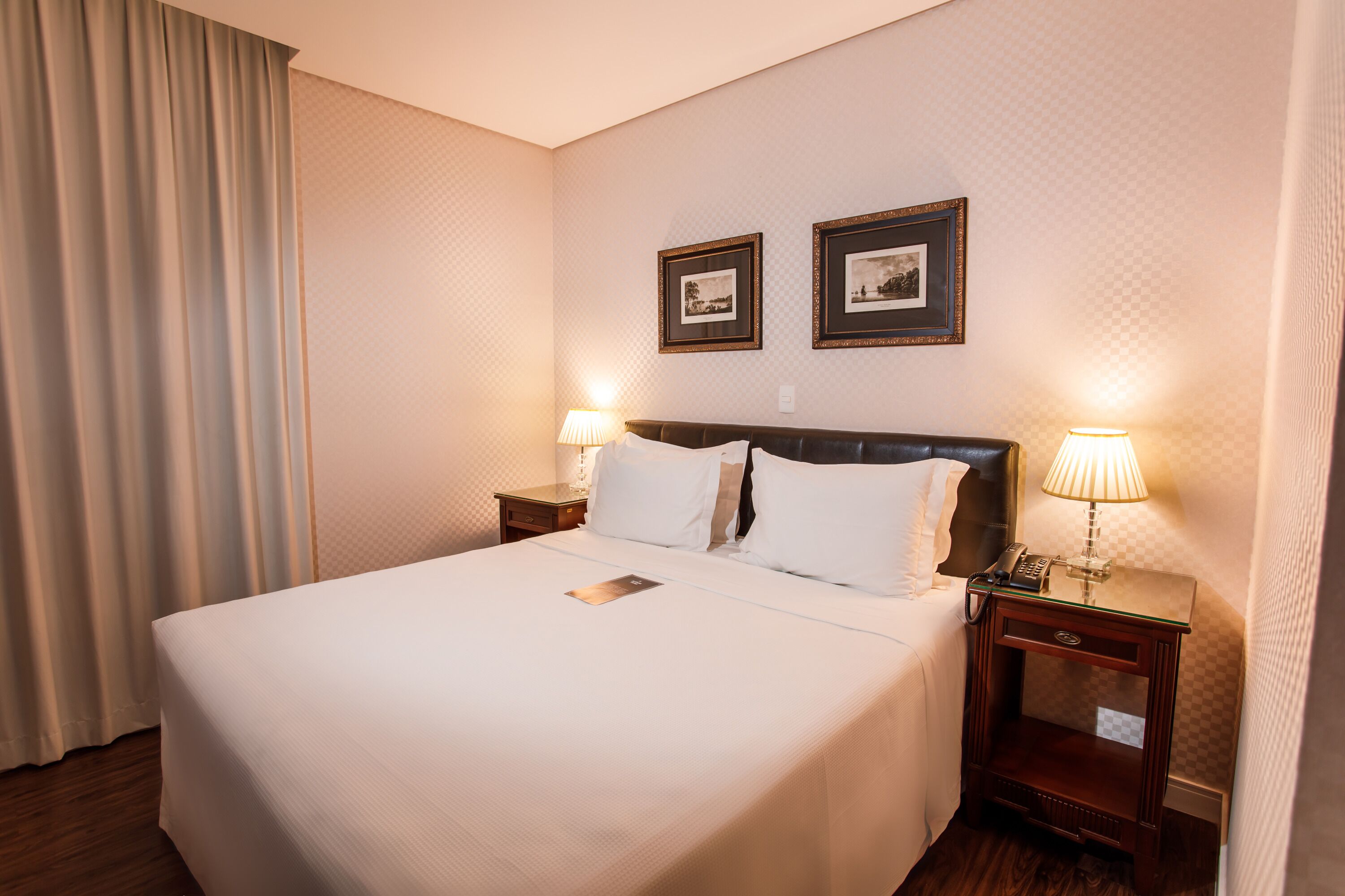 Quarto de hotel com uma cama grande, duas mesas de cabeceira com luminárias e duas fotos emolduradas na parede.