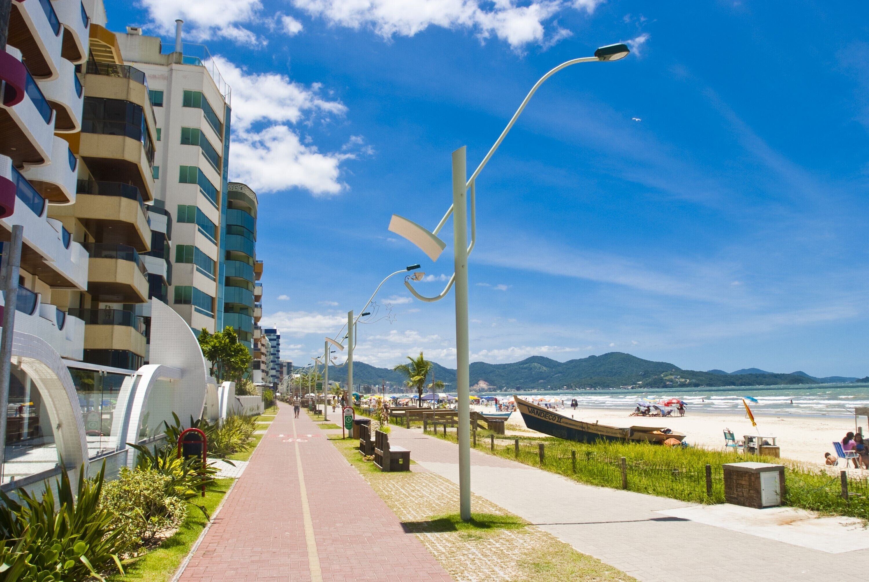 A coastal walkway lined with modern buildings, a beach with boats, and a clear blue sky.
