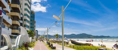 A coastal walkway lined with modern buildings, a beach with boats, and a clear blue sky.