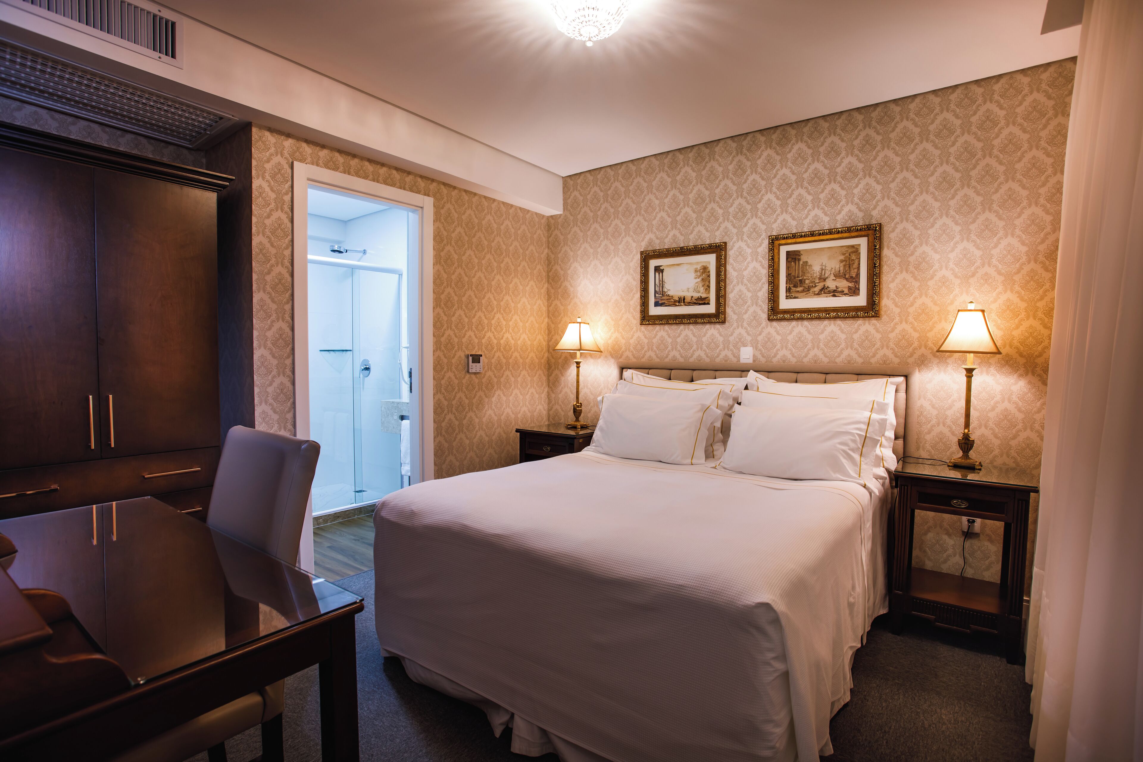 Habitación de hotel con cama, dos lámparas de noche, mesita de noche, armario de madera y escritorio con silla.