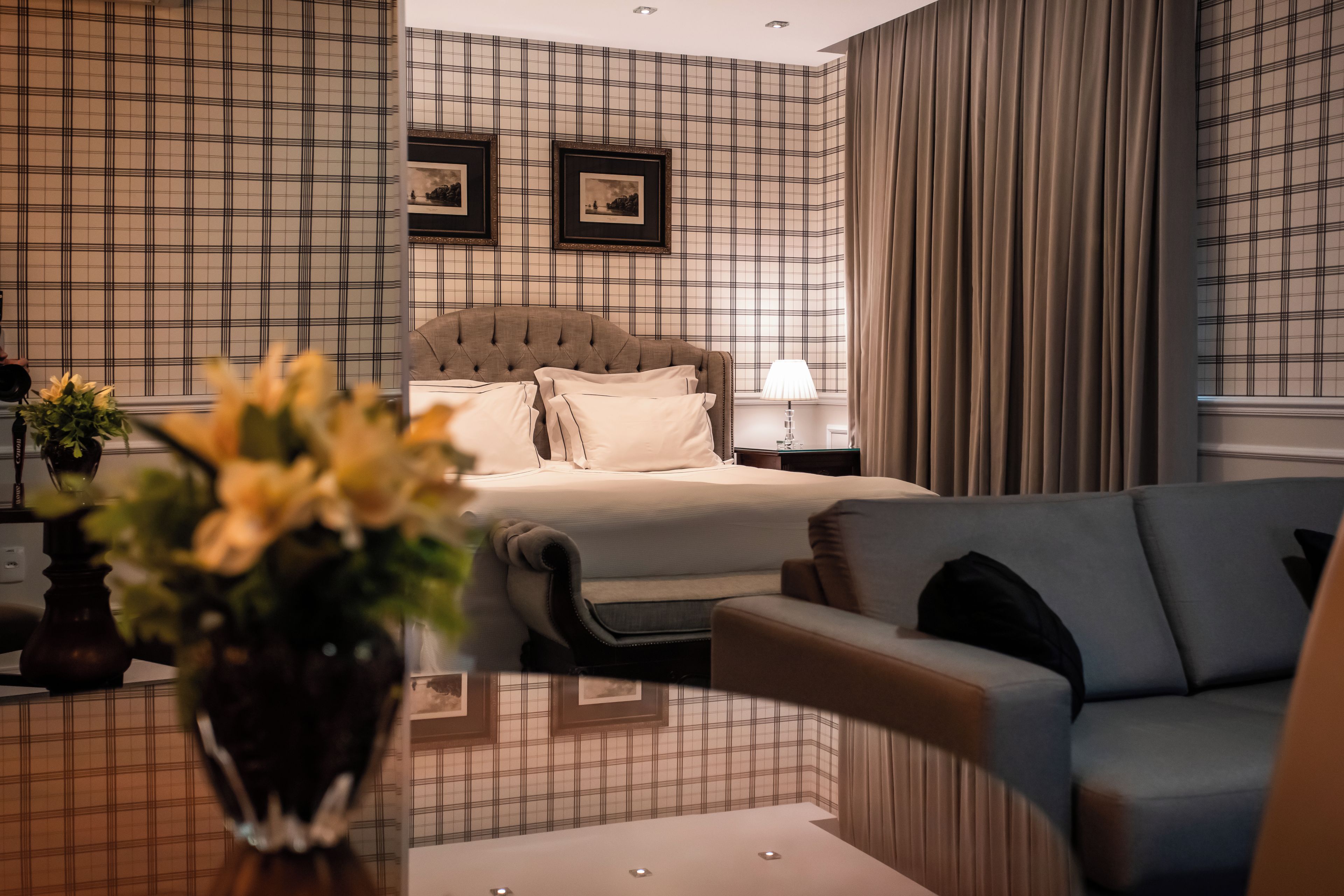 A hotel room with a bed, a nightstand, a lamp, a sofa, and a vase with flowers.