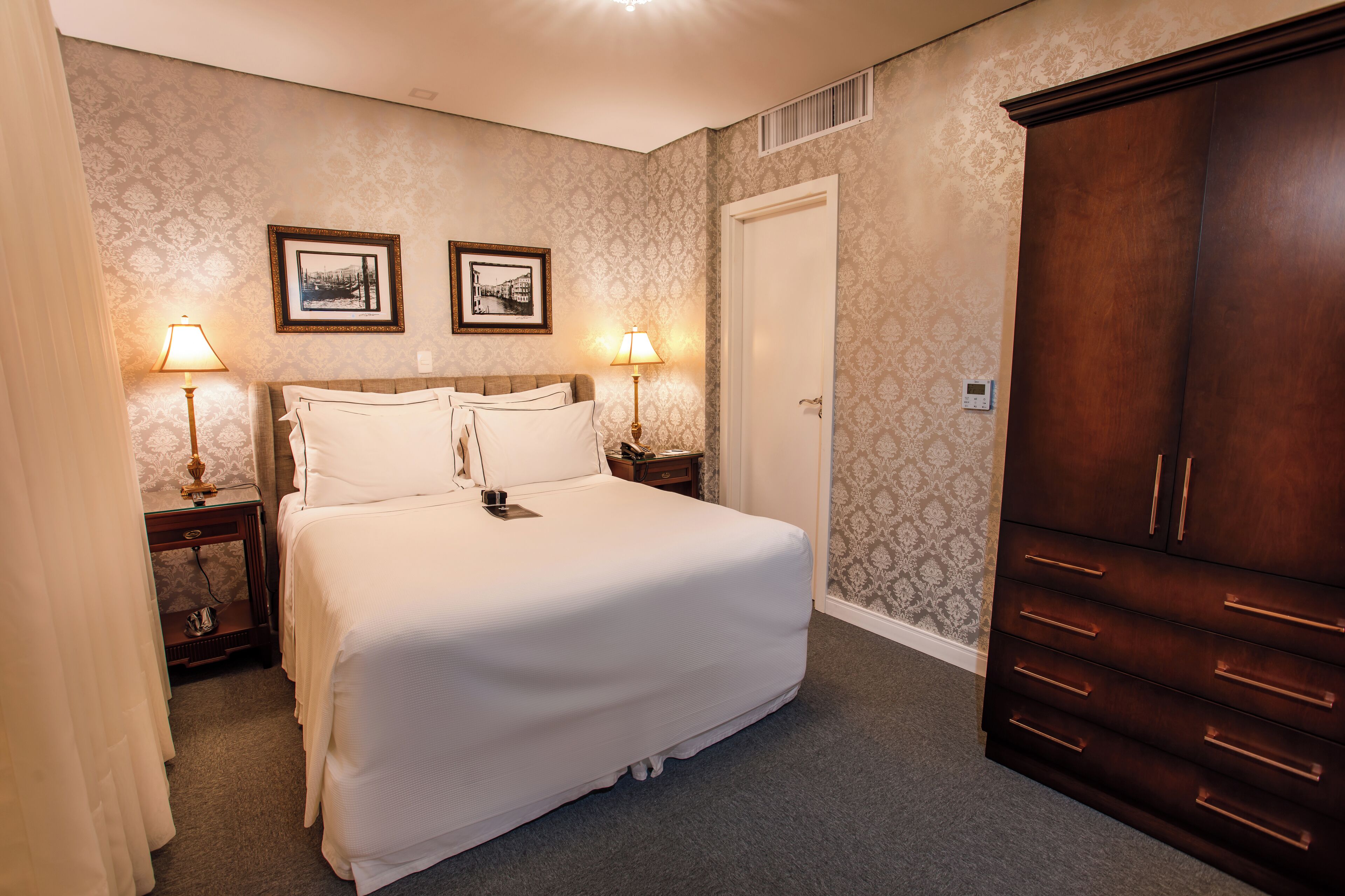 A hotel room with a large bed, two bedside tables with lamps, a wardrobe, and two framed pictures on the wall.