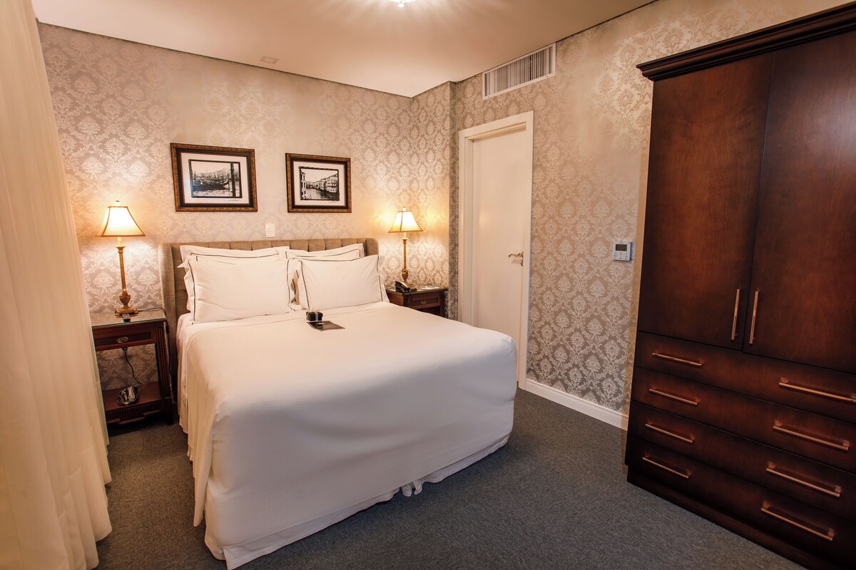 Habitación de hotel con una cama grande, dos mesitas de noche con lámparas, un armario y dos cuadros en la pared.