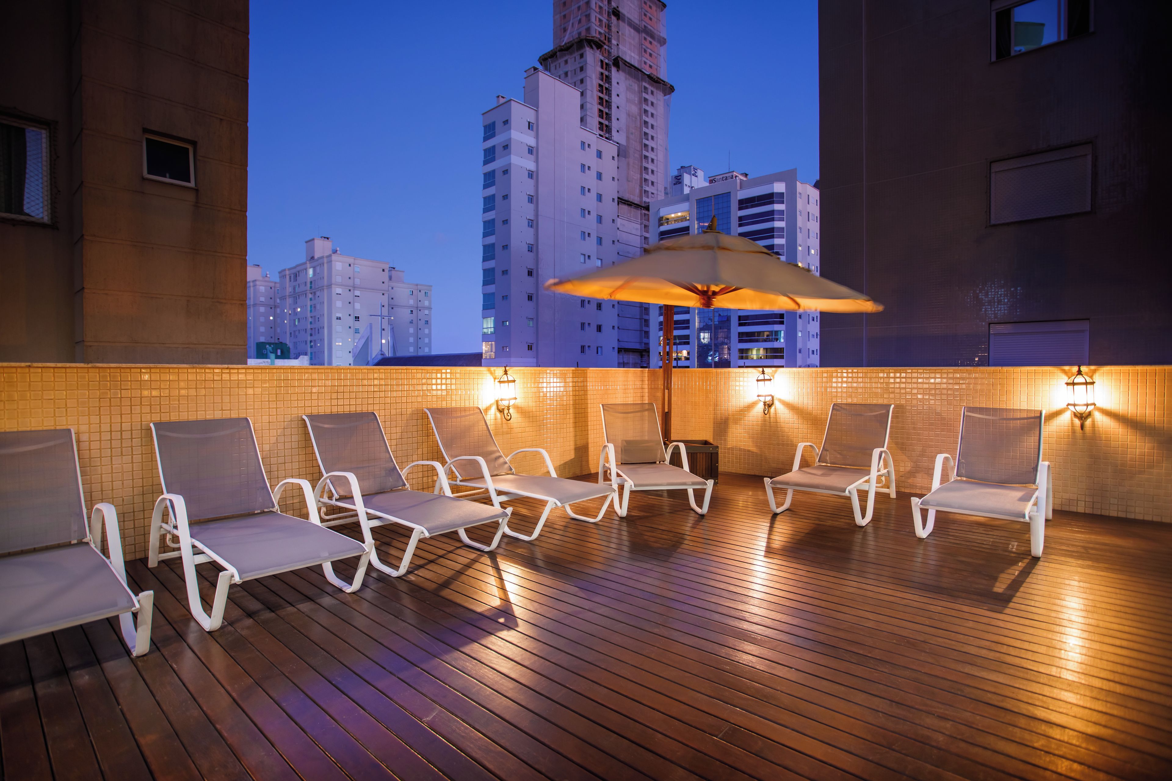 Um lounge no terraço com espreguiçadeiras e um grande guarda-sol, com vista para o horizonte da cidade ao entardecer.