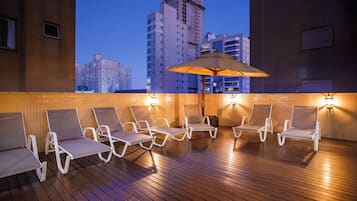 Um lounge no terraço com espreguiçadeiras e um grande guarda-sol, com vista para o horizonte da cidade ao entardecer.