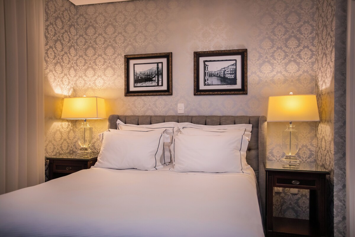 Une chambre d’hôtel avec un lit, deux tables de chevet avec des lampes et deux tableaux encadrés au mur.