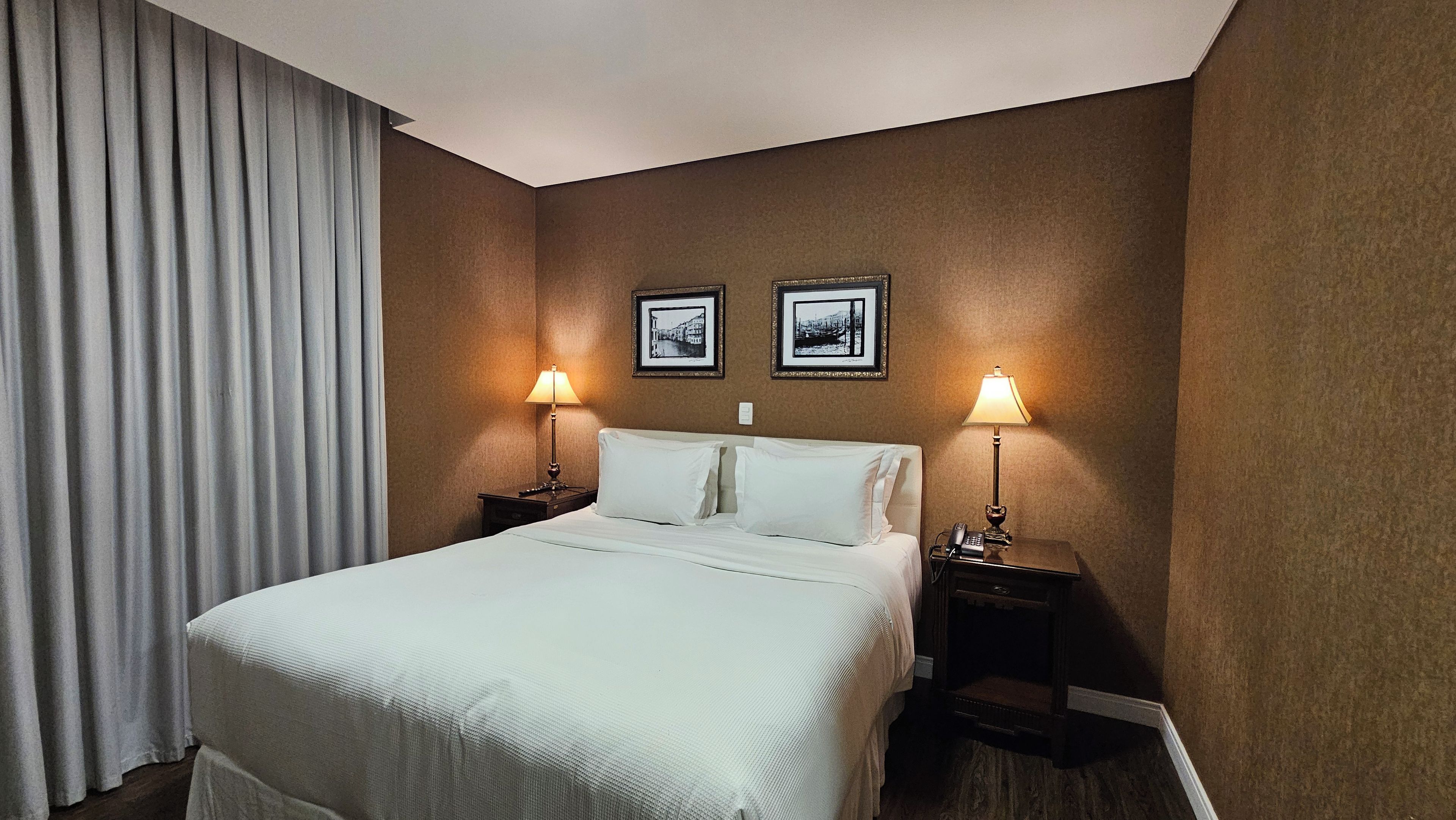 Une chambre d’hôtel avec un lit, deux lampes de chevet, une table de chevet et deux tableaux encadrés au mur.