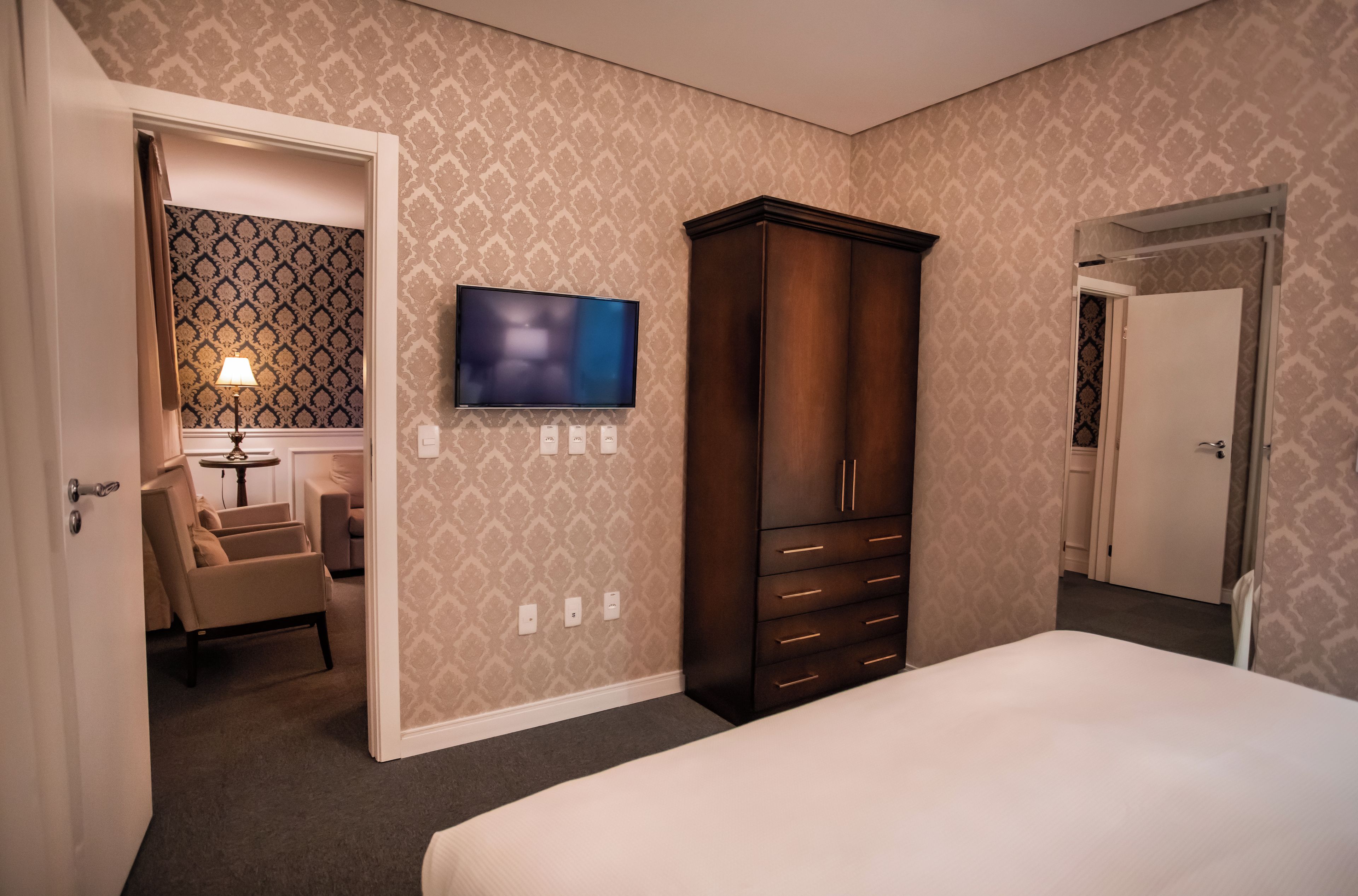 A hotel room with a bed, a television mounted on the wall, a wardrobe, and a desk with a lamp.