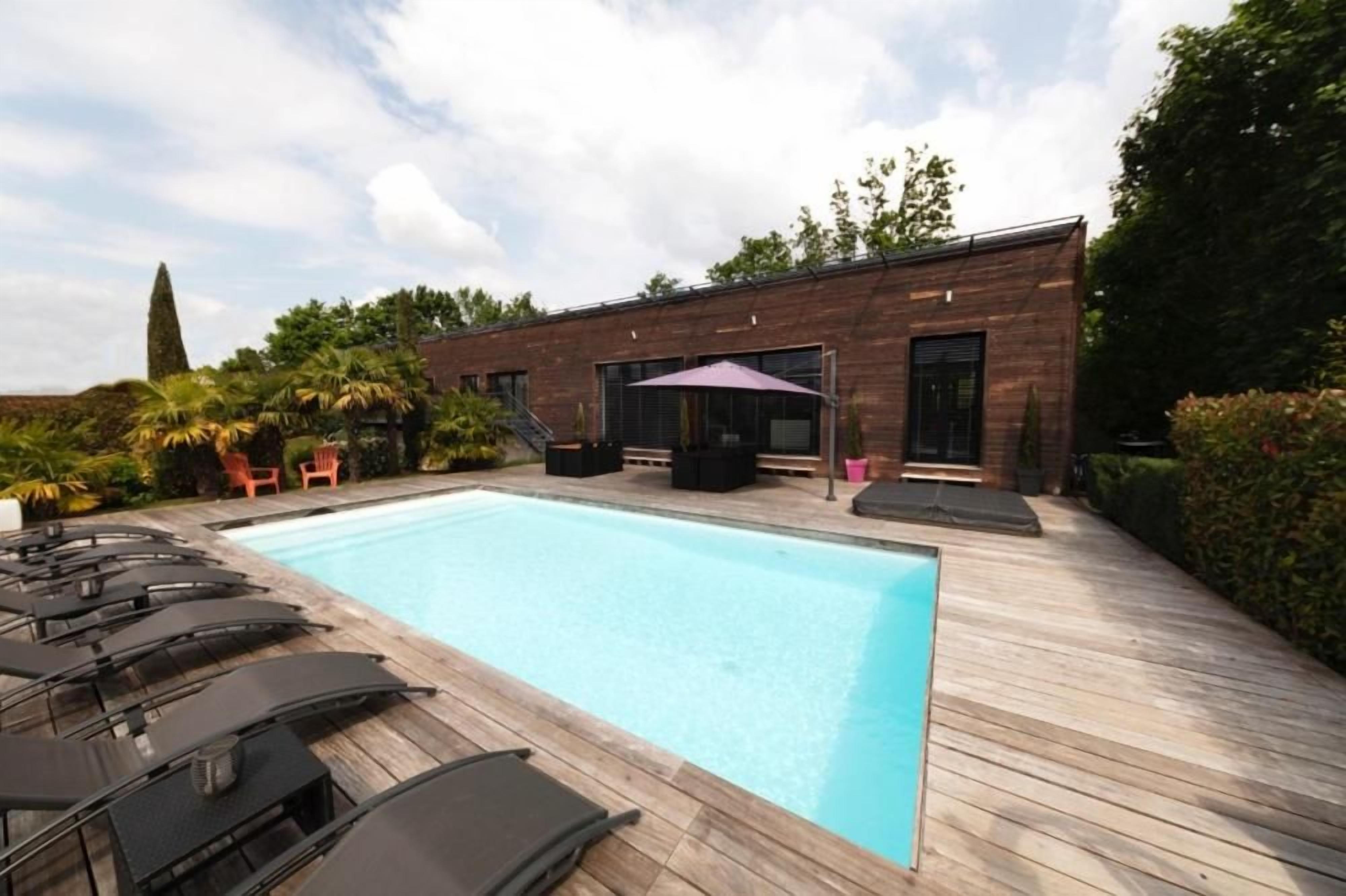 Una piscina con sillones de descanso, una terraza de madera y un edificio con ventanas grandes.