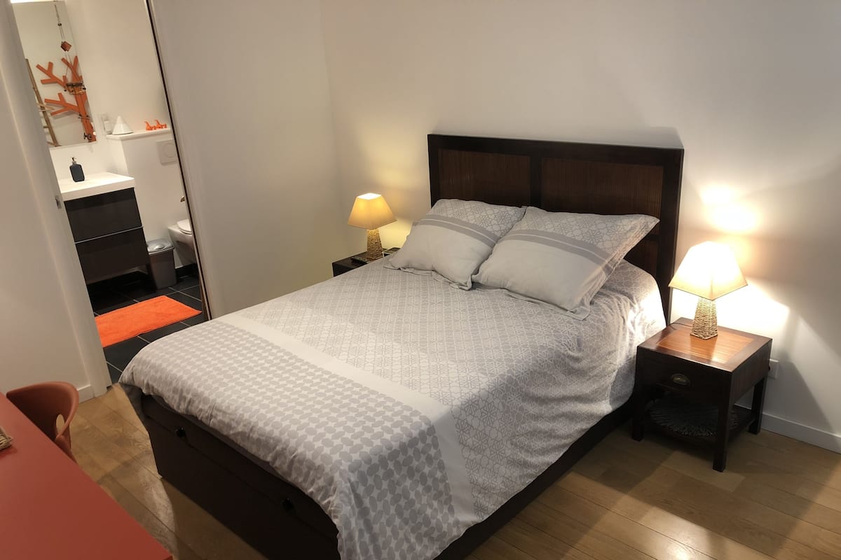 Una cama bien tendida con una colcha estampada y almohadas, flanqueada por dos mesitas de noche con lámparas, en una habitación con suelo de madera y un escritorio pequeño.