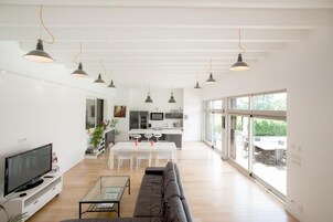 A modern living room with a sofa, coffee table, and TV, leading to a kitchen and dining area with large windows.