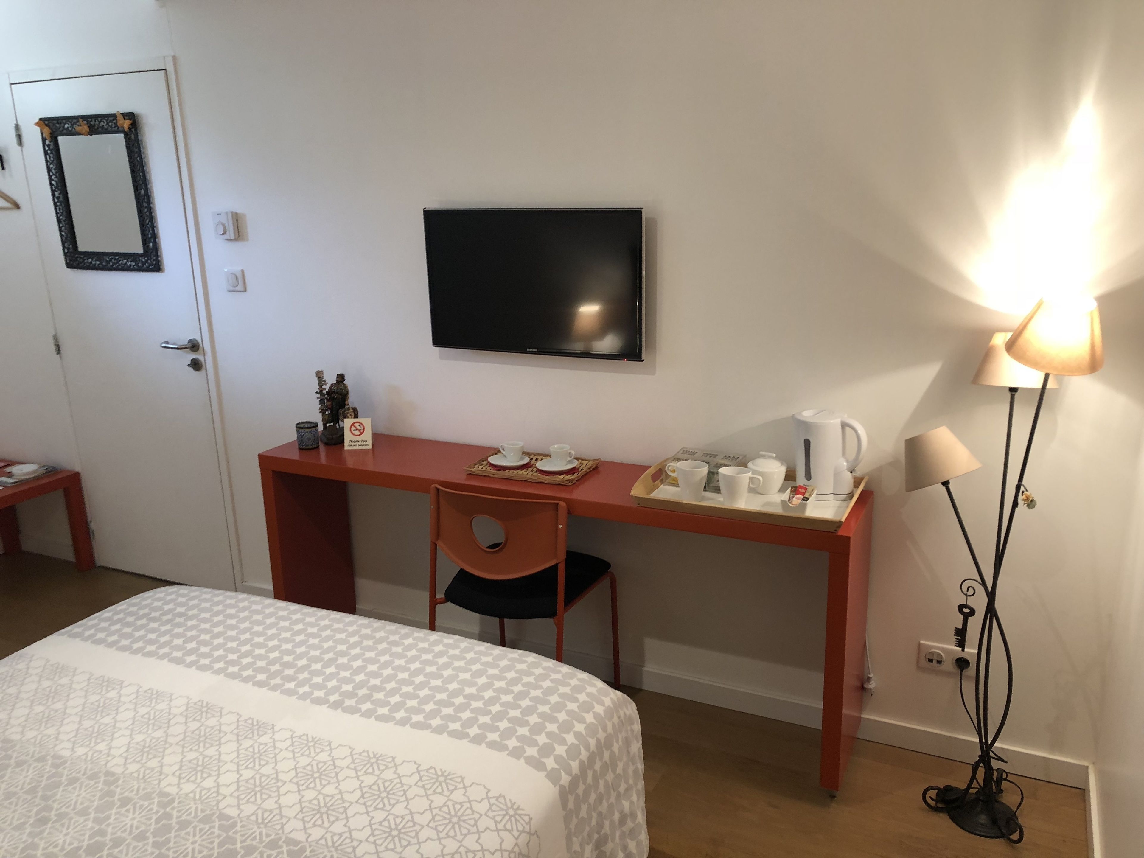 A hotel room with a bed, a desk with a chair, a television, a tray with cups and a kettle, and a floor lamp.