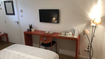 A hotel room with a bed, a desk with a chair, a television, a tray with cups and a kettle, and a floor lamp.