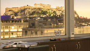 Une chambre d’hôtel avec vue sur un bâtiment historique perché sur une falaise, une fenêtre avec un rideau et une table sur laquelle se trouvent un journal et des verres.