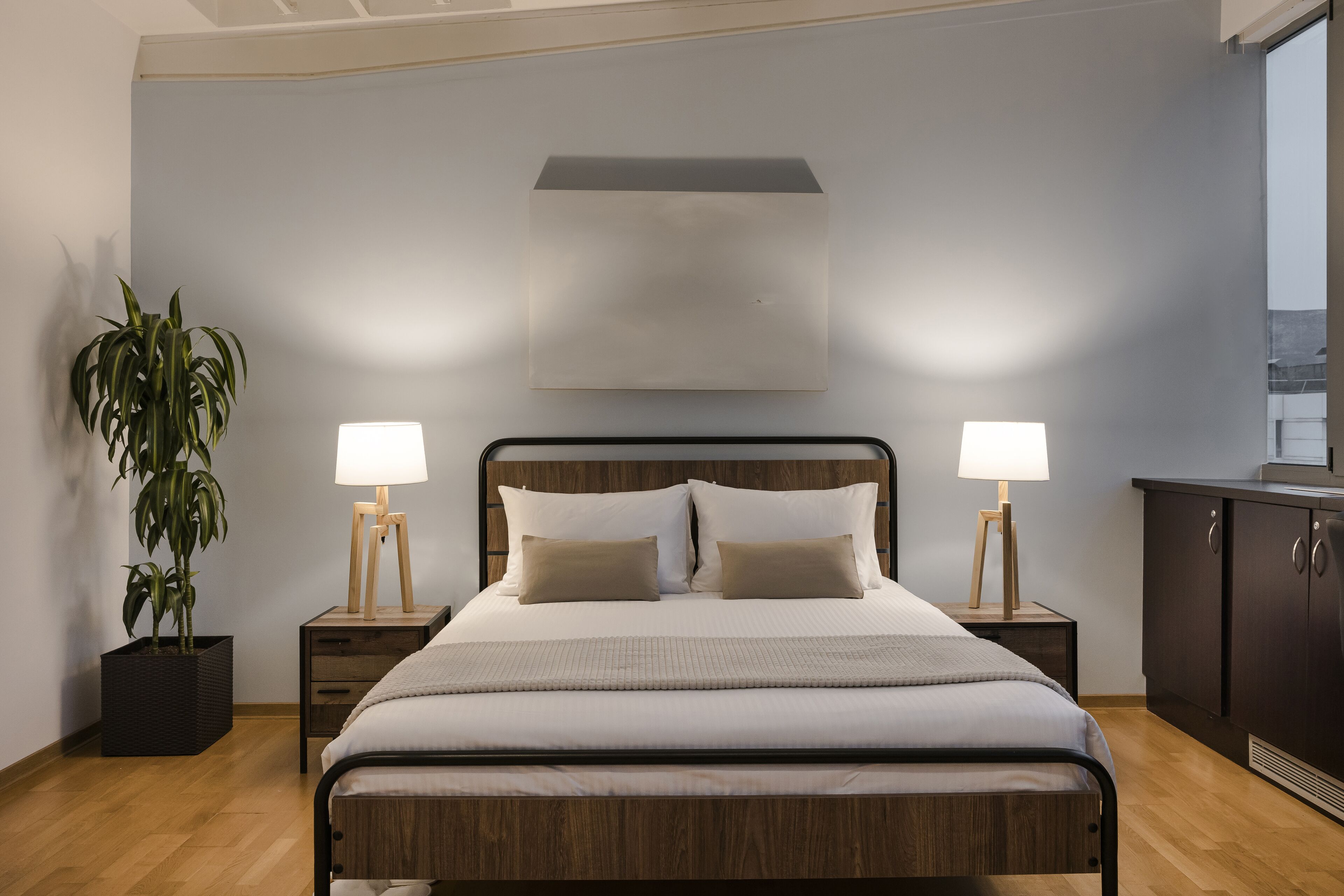 A neatly arranged bedroom with a bed, bedside lamps, a potted plant, and a cabinet.