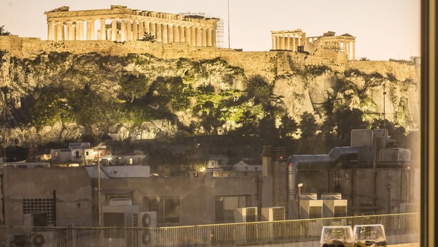 Vue sur l’Acropole d’Athènes depuis la fenêtre d’une chambre d’hôtel, avec deux verres de vin et une assiette de nourriture posés sur le rebord de la fenêtre.